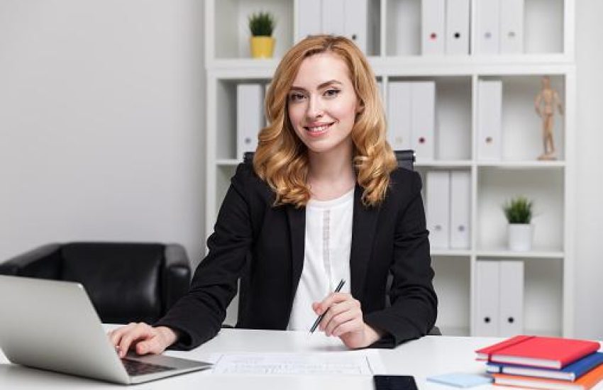 Smiling business lady sitting at her desk smiling and holding pen. Hand on laptop keyboard. Concept of busy manager.